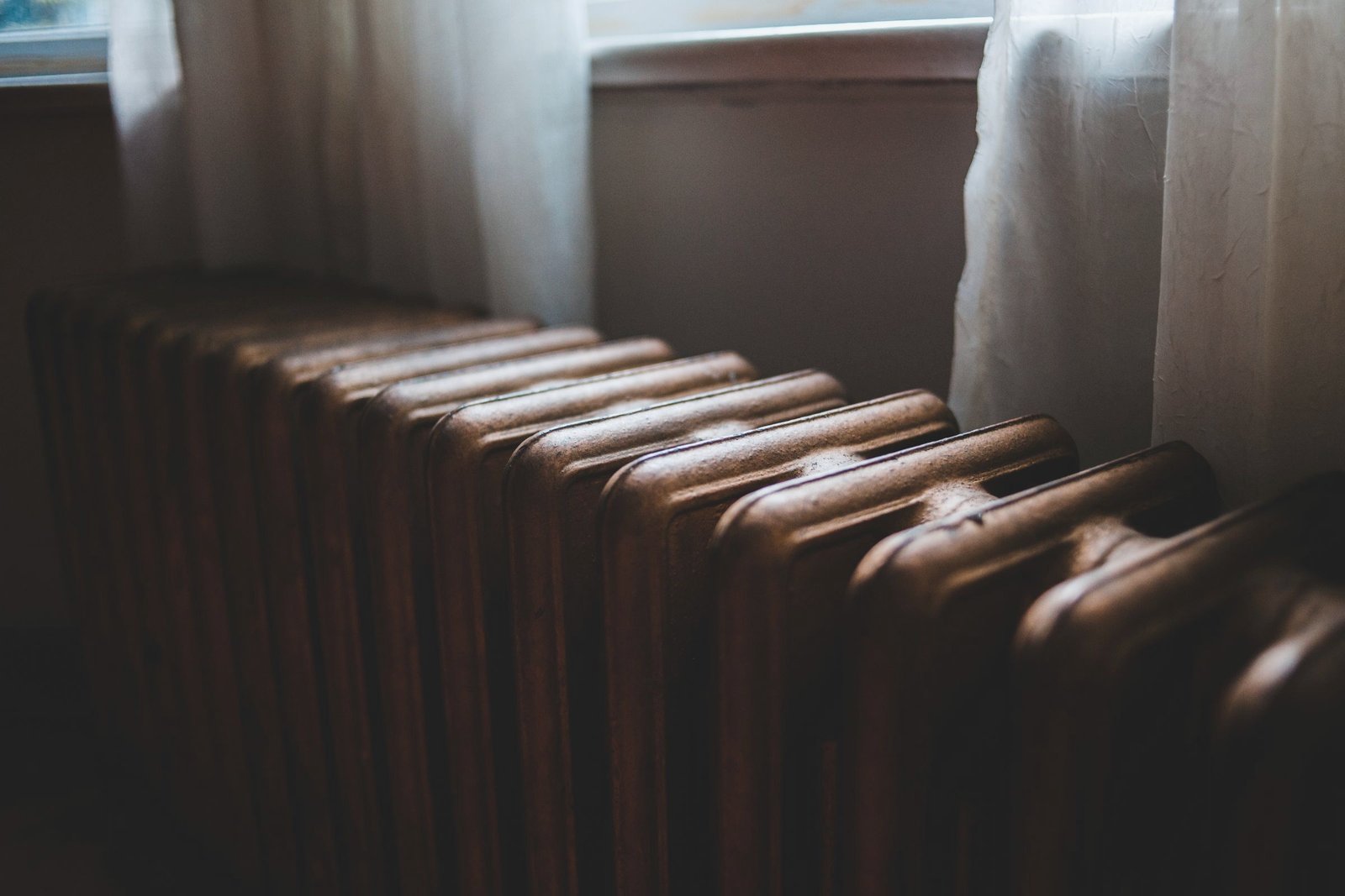 A cast iron radiator below a window with sheer curtains, in low winter light.
