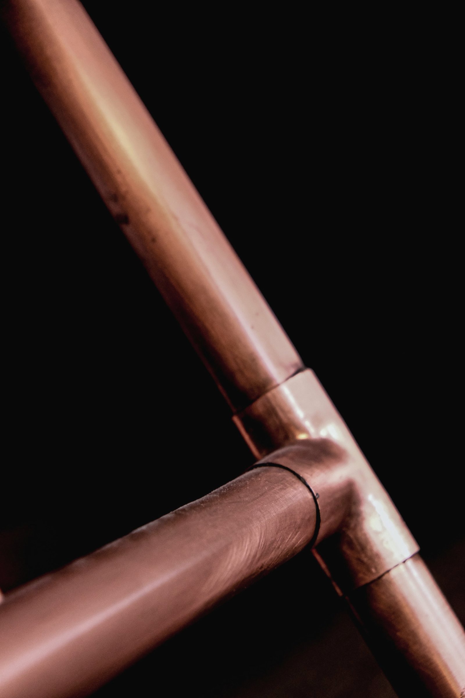 Warm-lit copper supply pipes meeting at a soldered tee joint against a dark wall.