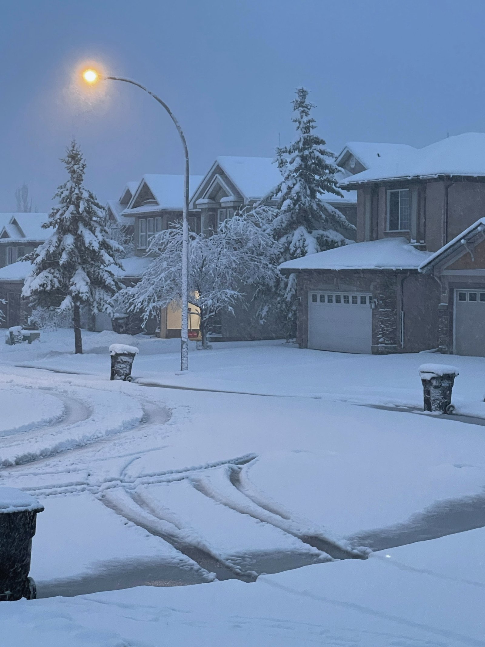 A residential street under deep snow at dusk, one streetlight glowing warm.